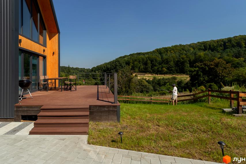 Котеджне містечко КМ Схили: двоповерховий будинок з дерев'яною обшивкою та терасою, прибудинкова територія з газоном та огорожею.