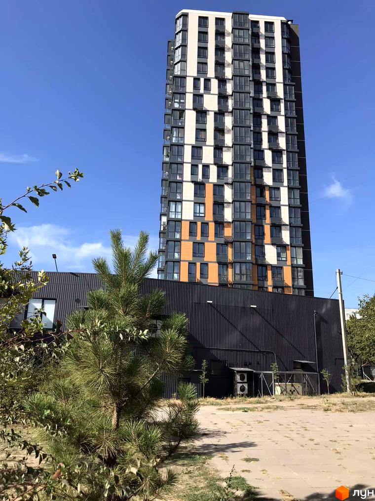 Житловий комплекс River Hall – висотна новобудова з панорамними вікнами та оранжевими вставками. Навколо будівлі розташована прибудинкова територія з деревами та асфальтованим покриттям.