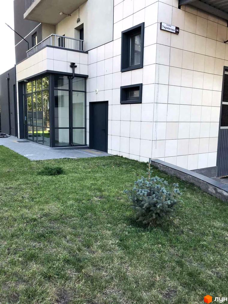 Зовнішній вигляд новобудови ЖК River Hall з білою плиткою та темними вікнами, зелена галявина з молодим деревом перед фасадом.