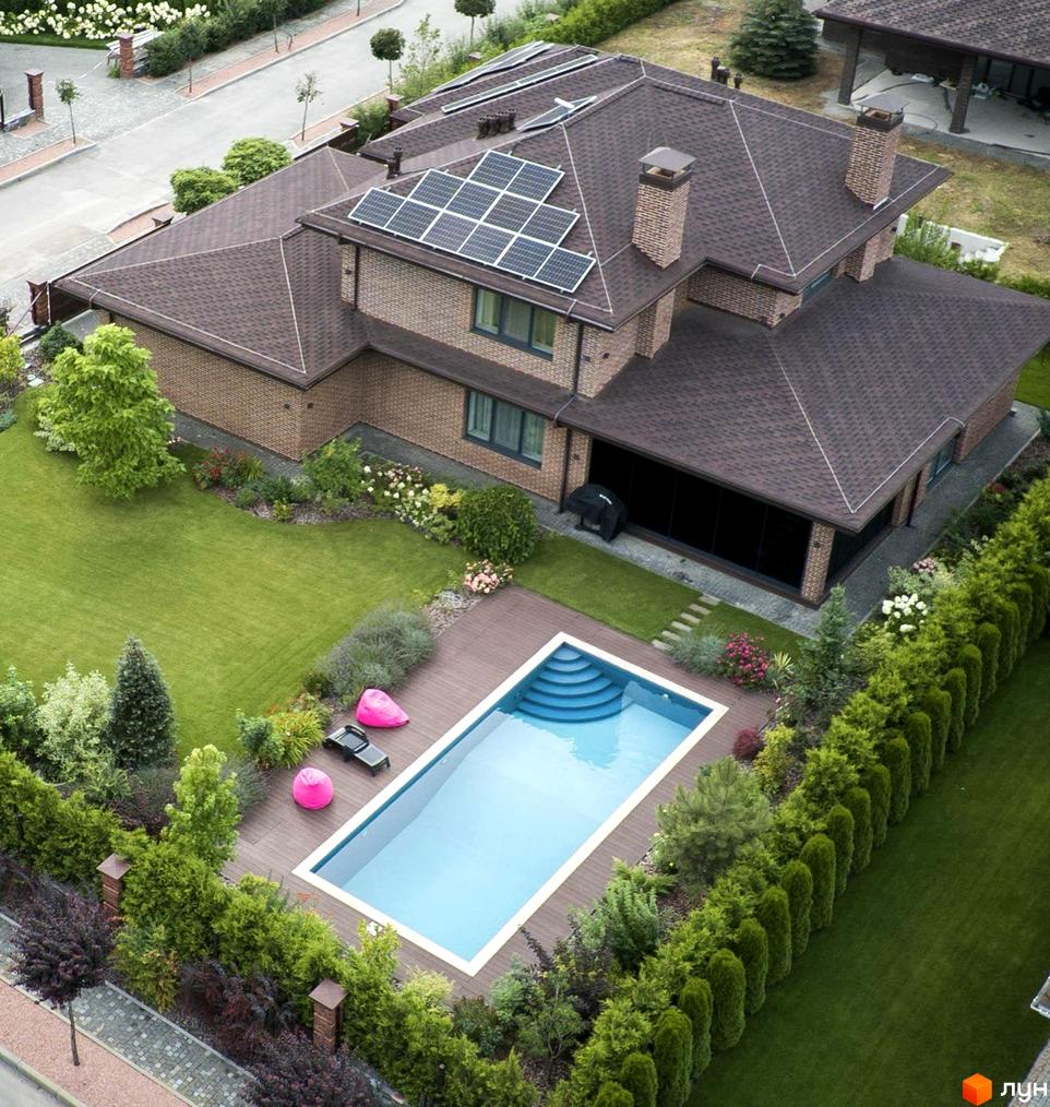 Котеджне містечко Вітаград: двоповерховий будинок з коричневим дахом, сонячними панелями та басейном на задньому дворі. Навколо будинку доглянутий газон та зелені насадження.