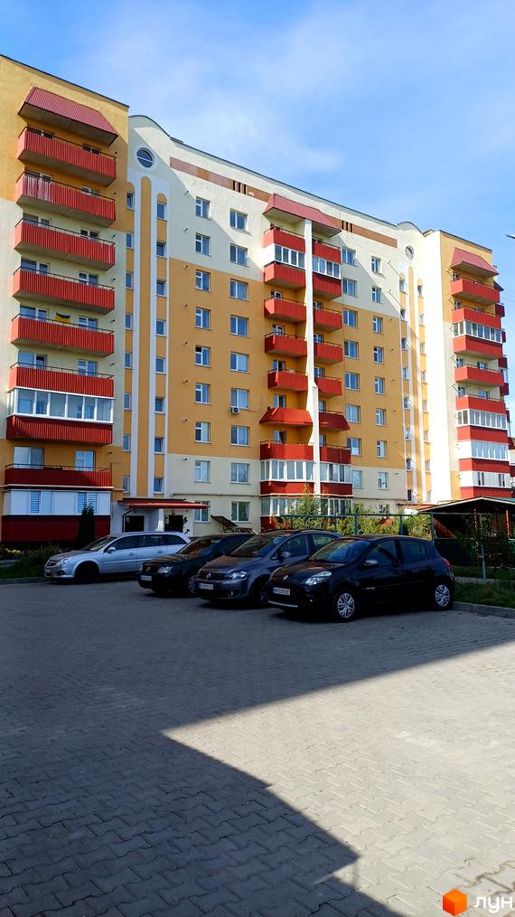 Зображення фасаду багатоповерхового будинку на вулиці Трудова, 7/9. Перед будинком припарковані автомобілі на брукованій парковці.