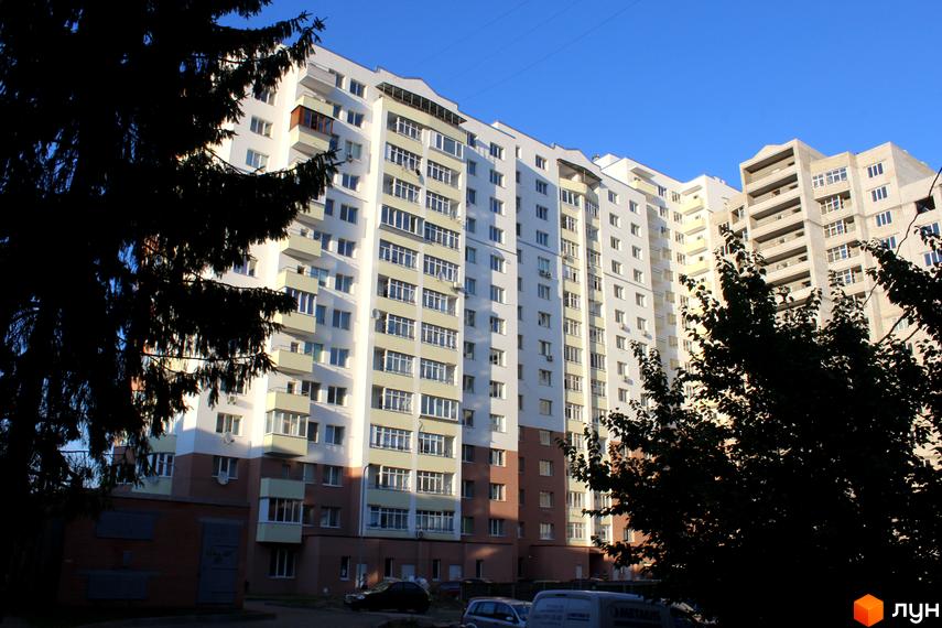 Житловий комплекс Княжа гора: багатоповерховий будинок білого кольору зі світлими балконами та коричневою основою. Навколо видно дерева та автомобілі.