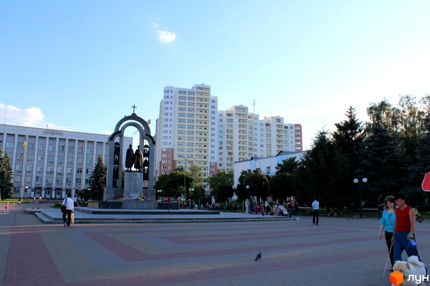 Багатоповерховий житловий комплекс Княжа гора з білим фасадом та балконами. Перед будинком розташована простора площа з пам'ятником, деревами та людьми.