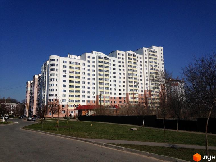 Багатоповерховий житловий комплекс Княжа гора з білим фасадом та світлими балконами. На передньому плані дорога, зелений газон та дерева без листя.