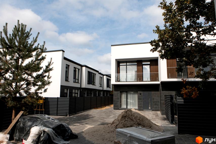 Котеджне містечко KМ Balance Homes: двоповерхові будинки з білими фасадами та темними елементами, балкон з скляним огородженням. Прибудинкова територія з бруківкою та чорним парканом.