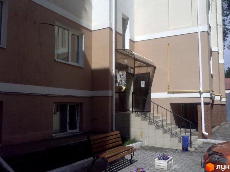 Фасад житлового комплексу Авангард на „Лісовій“ у світло-коричневих тонах з білими акцентами. На прибудинковій території видно вхід зі сходами, лавку та бруківку.