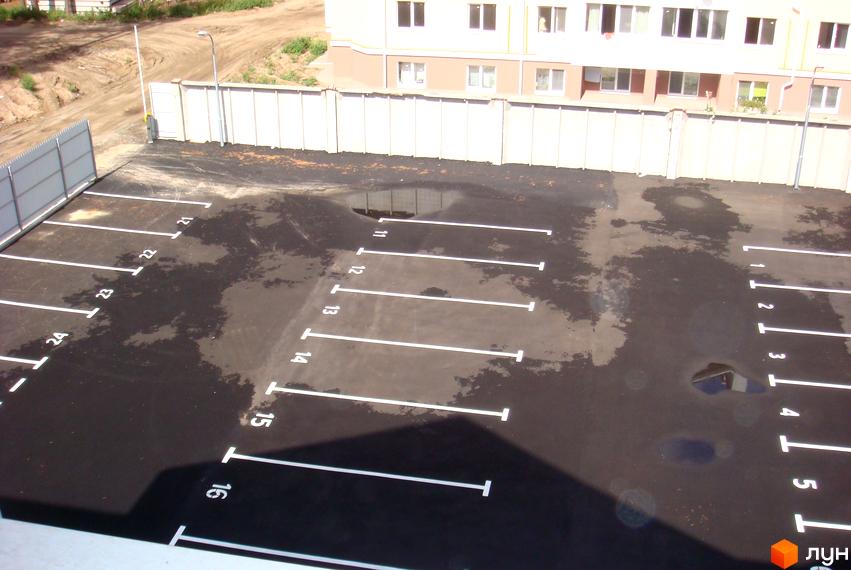 Фасад будинку житлового комплексу Авангард на „Лісовій“ світлого кольору з коричневою основою. Перед будинком розташований асфальтований паркінг з розміткою та номерами місць.