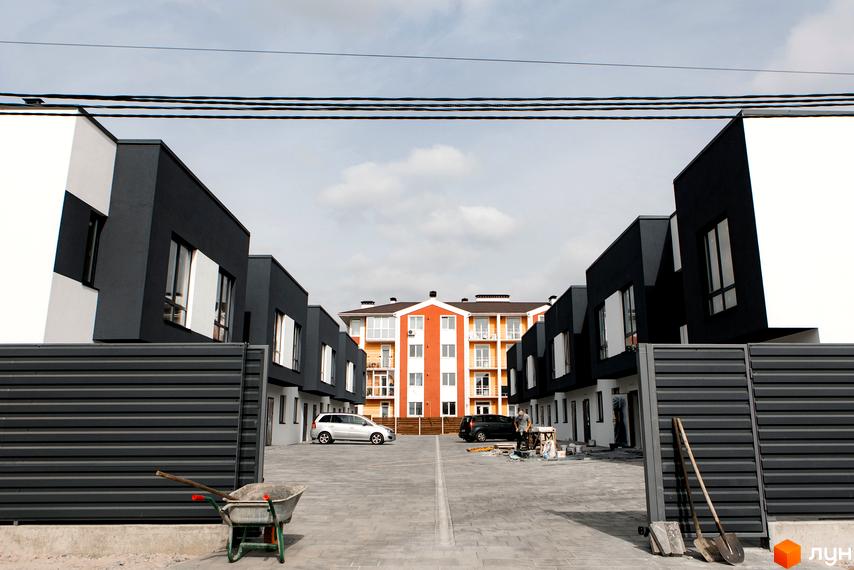 На фото Таунхауси SDG Bilohorodka з темно-сірими та білими фасадами, а також багатоповерховий будинок. Прибудинкова територія вимощена бруківкою, видно припарковані автомобілі.