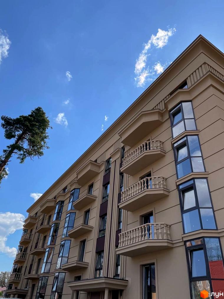 Фасад житлового комплексу Monaco з балконами та великими вікнами на тлі блакитного неба з хмарами. Навколо будівлі видно зелені дерева.