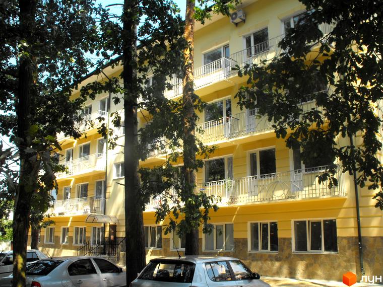 Зовнішній вигляд чотириповерхового будинку з балконами. На передньому плані дерева та автомобілі.