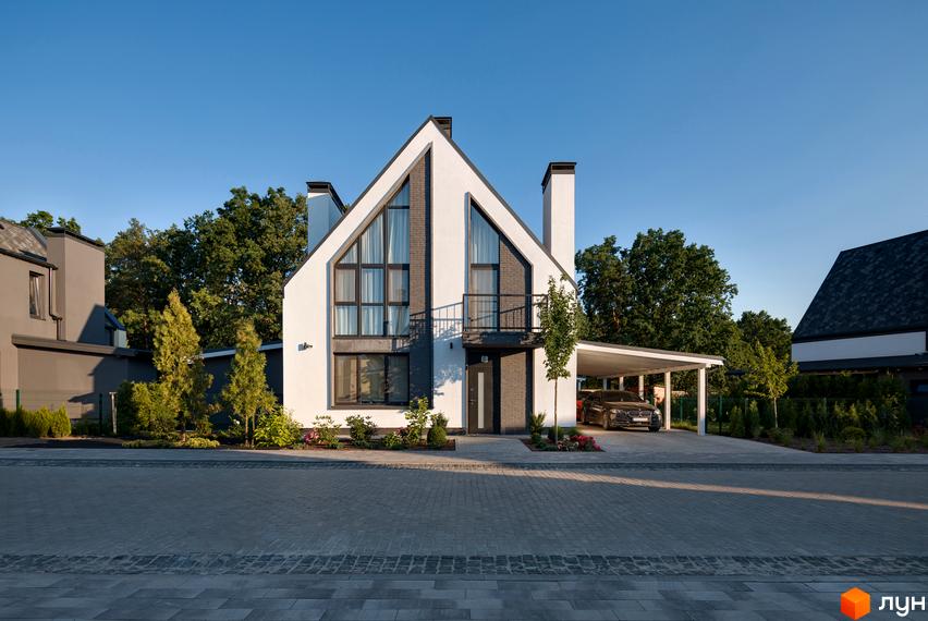 Котеджне містечко KM VILLAGGO. Сучасний двоповерховий будинок з білою фасадною штукатуркою, великими панорамними вікнами та навісом для автомобіля. Навколо будинку доглянутий ландшафт з деревами та кущами.