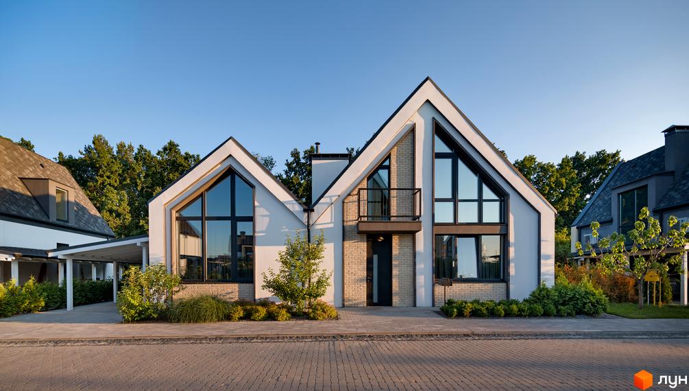 Котеджне містечко КМ VILLAGGO. Сучасний будинок з великими вікнами та двосхилим дахом, оточений зеленню та вимощеною доріжкою.