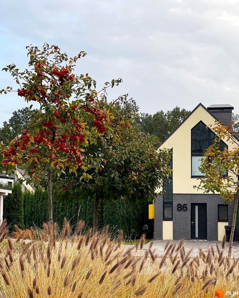 Екстер'єр будинку №86 в котеджному містечку КМ VILLAGGO. Навколо будинку розташовані дерева з червоними плодами та декоративні трави.