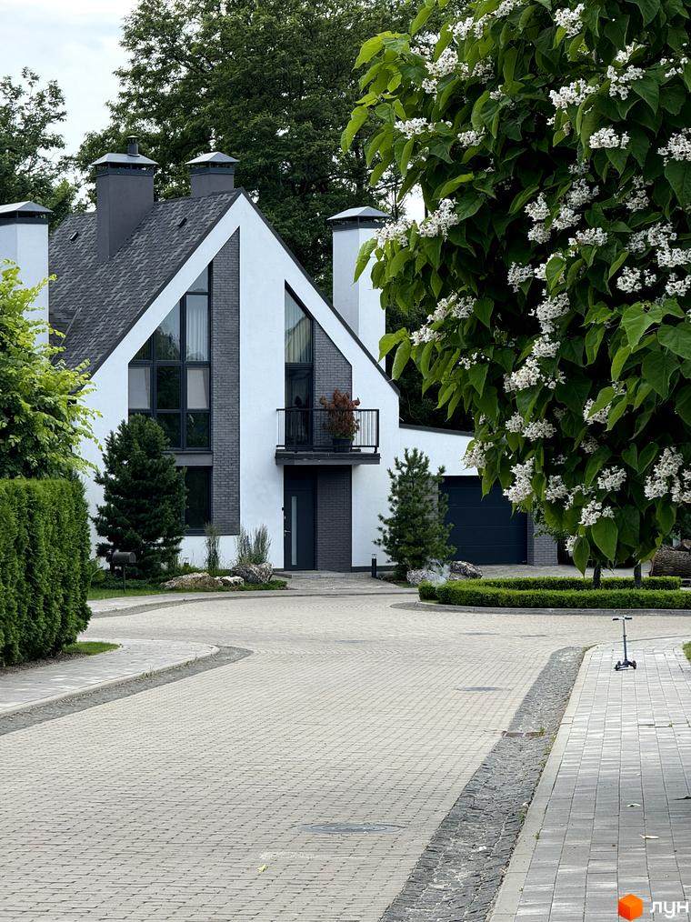 Котеджне містечко KM VILLAGGO. Сучасний будинок з великими вікнами та двома димарями на тлі зелені та квітучого дерева. Доглянута прибудинкова територія з бруківкою та кущами.