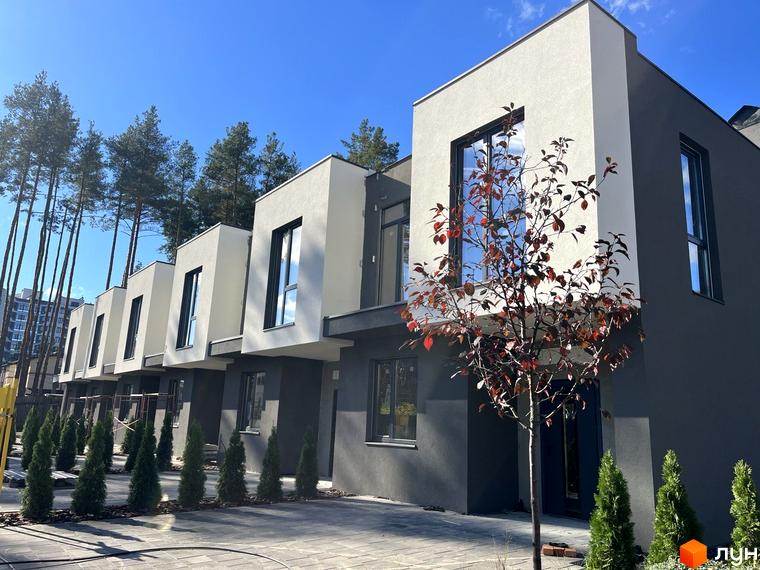 Ряд двоповерхових будинків з фасадами світлого та темного сірого кольору. Прибудинкова територія облаштована бруківкою та молодими хвойними деревами.