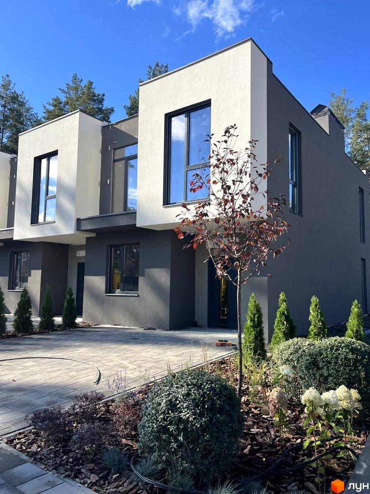 Будинок з сірим та білим фасадом та великими вікнами. Прибудинкова територія з зеленими насадженнями та бруківкою.