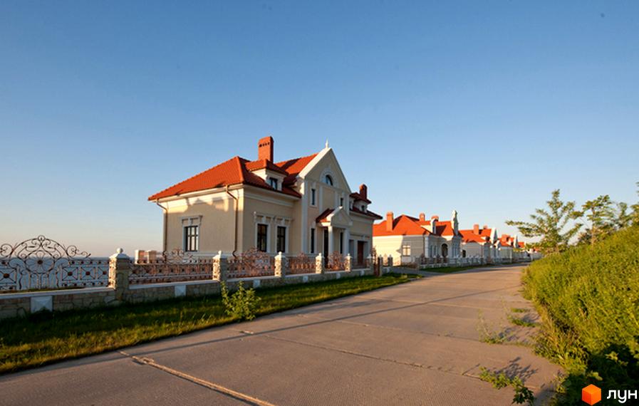 Фото Котеджного містечка Сосновий берег. Двоповерховий будинок з червоним дахом та світлим фасадом, перед будинком газон та кована огорожа.