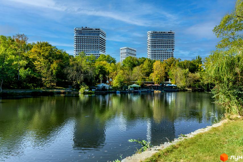 Житловий комплекс White Lines на березі озера. Три високі будинки з панорамними вікнами, зелена територія з деревами та водою.