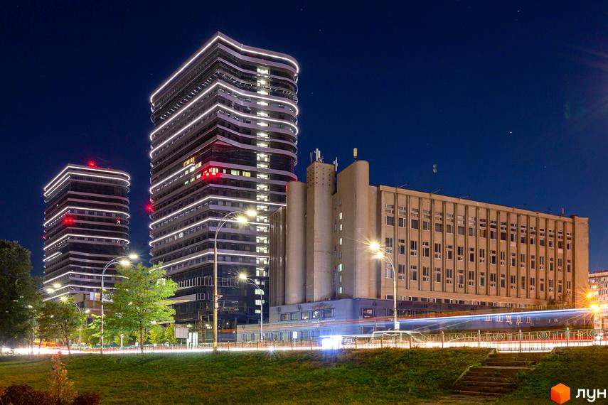 Житловий комплекс White Lines вночі: дві висотні будівлі з вигнутим фасадом та горизонтальними білими світловими лініями, поруч дорога та зелені насадження.