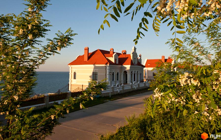 Зображення Котеджного містечка Сосновий берег з червоним дахом та видом на море. На передньому плані зелені дерева та доглянута територія.