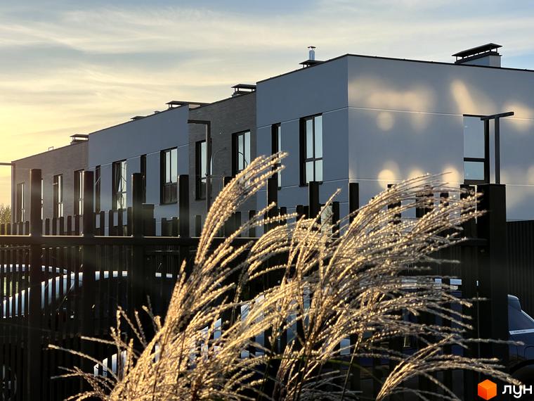 Ряд таунхаусів Soul City зі світлими фасадами та темними вікнами під блакитним небом. На передньому плані видно металевий паркан та освітлені сонцем рослини.