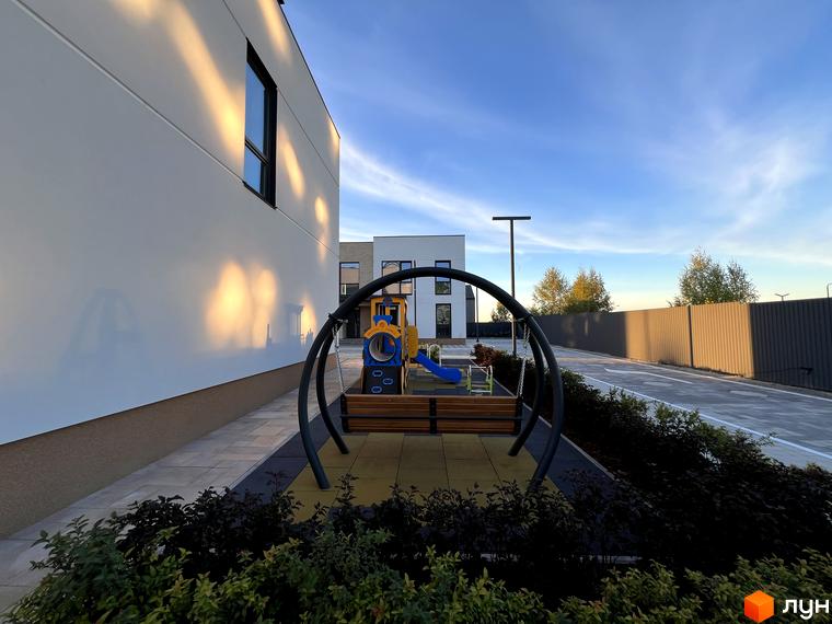 Фасад таунхаусу Soul City білого кольору з темними вікнами, поруч дитячий майданчик з гойдалкою та гіркою.