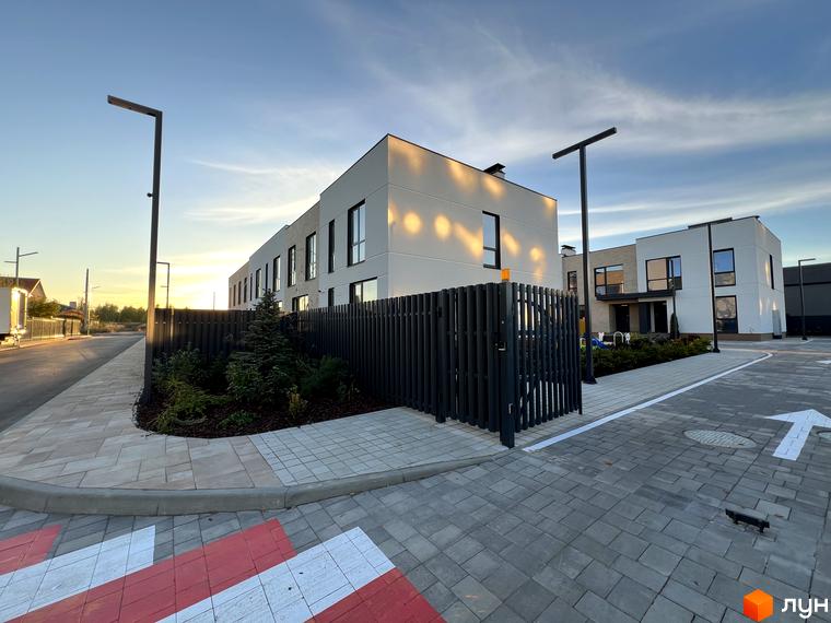 Фасади Таунхаусів Soul City світлого кольору з темними вікнами. На прибудинковій території дорога з бруківки, огорожа та зелені насадження.