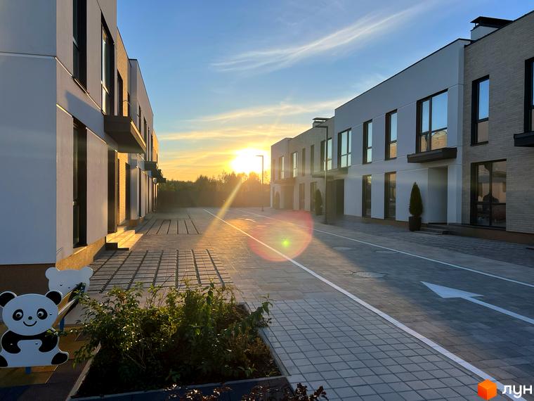 Таунхауси Soul City з фасадами білого та сірого кольору на тлі заходу сонця. Поруч розташована вимощена бруківкою територія та зелені насадження.