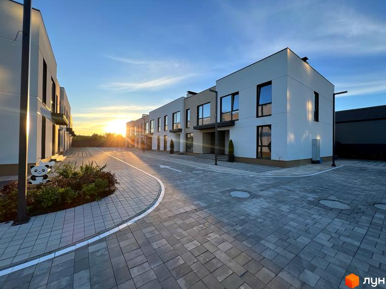 Таунхауси Soul City з білими фасадами та темними вікнами, розташовані на бруківці з висадженими рослинами. Навколо території видно дерева та блакитне небо з сонцем.