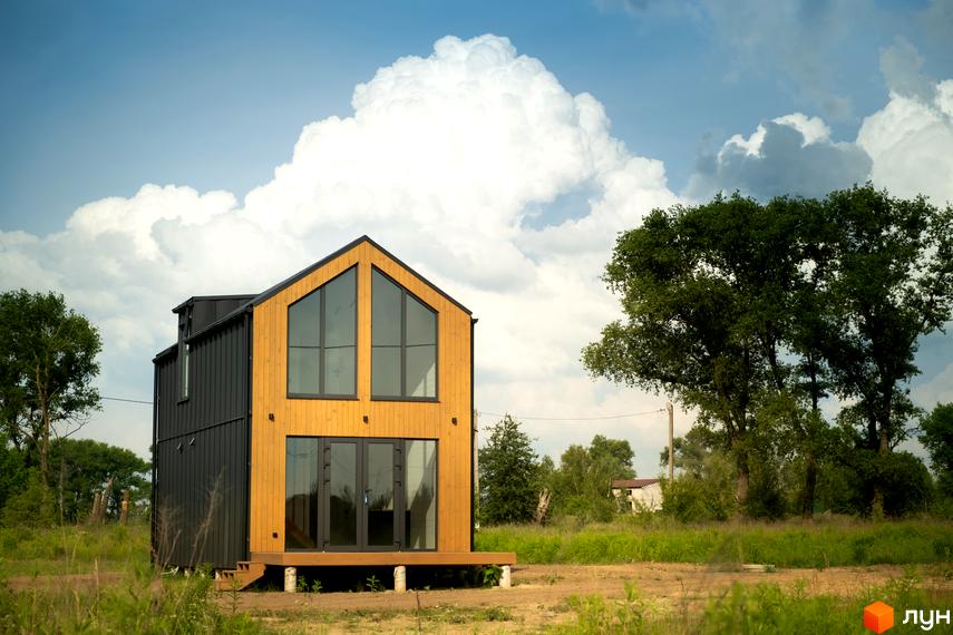 Котеджне містечко Залісся Village: двоповерховий будинок з дерев'яним фасадом та великими вікнами на тлі хмарного неба. Навколо зелена трава та дерева.