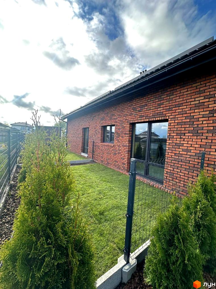 Вигляд ззовні цегляного будинку в Котеджному містечку Одноповерховий Київ - Зубра. Навколо будинку зелений газон та декоративні туї.