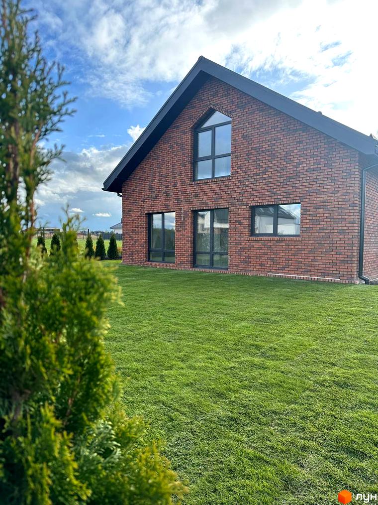 Фасад будинку з червоної цегли та чорними вікнами в Котеджному містечку Одноповерховий Київ - Зубра. Перед ним великий зелений газон та хвойні дерева.