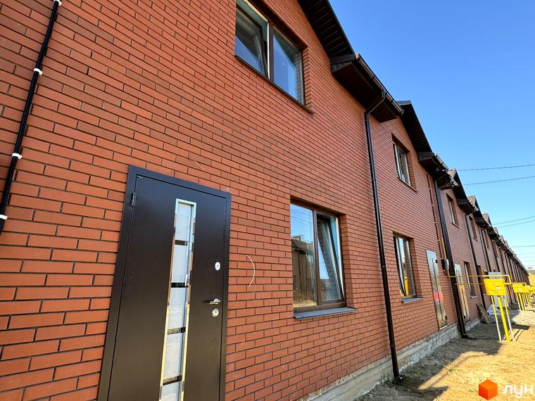 Котеджне містечко Весняний: фасади двоповерхових будинків з червоної цегли, темними вікнами та дверима, поруч неозеленена ділянка.