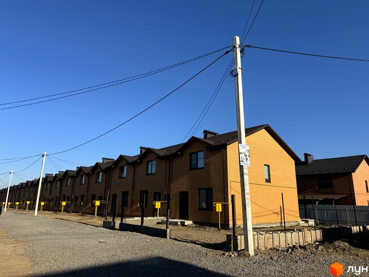 Котеджне містечко Весняний: ряд двоповерхових будинків з жовтої цегли та темними дахами. Перед будинками розташована гравійна дорога та стовпи електропередач.