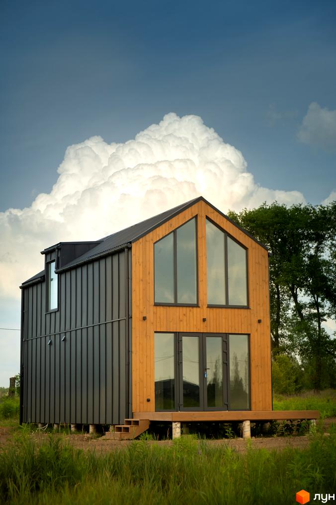 Котеджне містечко Залісся Village: двоповерховий будинок з дерев'яним фасадом та великими вікнами на тлі хмарного неба. Навколо будинку зелена трава та дерева.