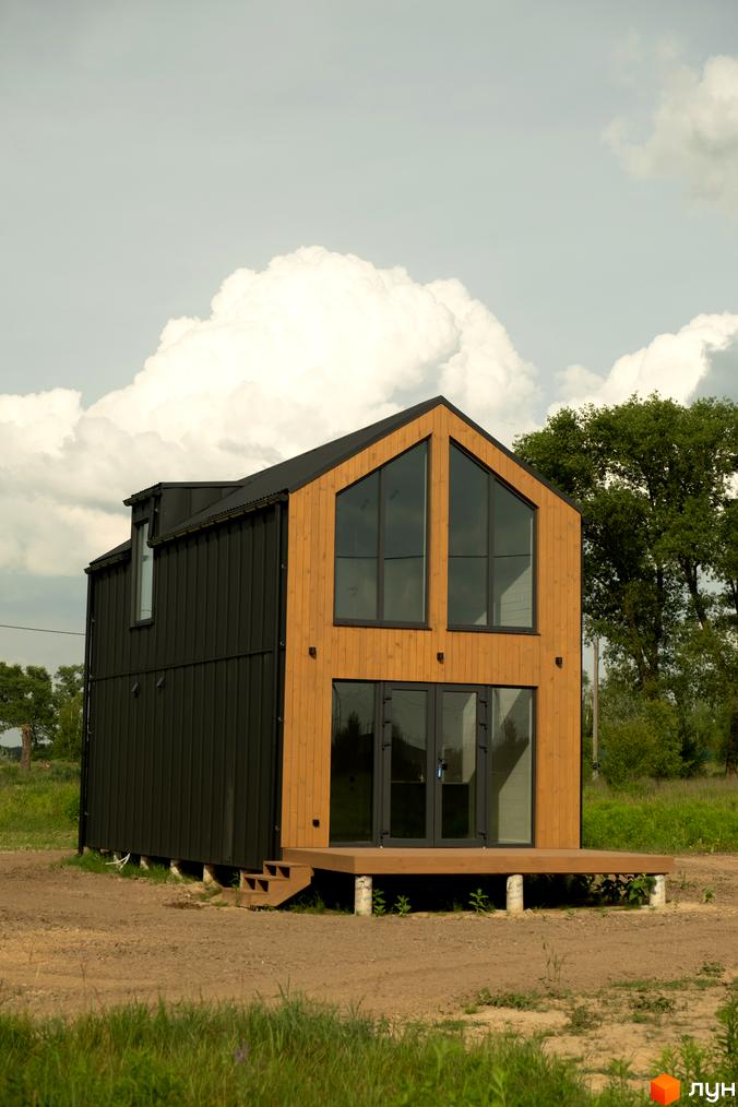 Двоповерховий будинок з чорним та дерев'яним фасадом, великими вікнами та терасою. Навколо будинку зелена трава та дерева.