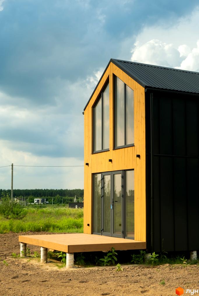 Котеджне містечко Залісся Village: двоповерховий будинок з дерев'яним фасадом та чорною металевою обшивкою, великими вікнами та терасою.