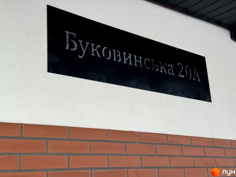 Фасад житлового комплексу Буковинська 20А, оздоблений білою штукатуркою та цегляною кладкою. На фасаді розміщена чорна вивіска з назвою комплексу.