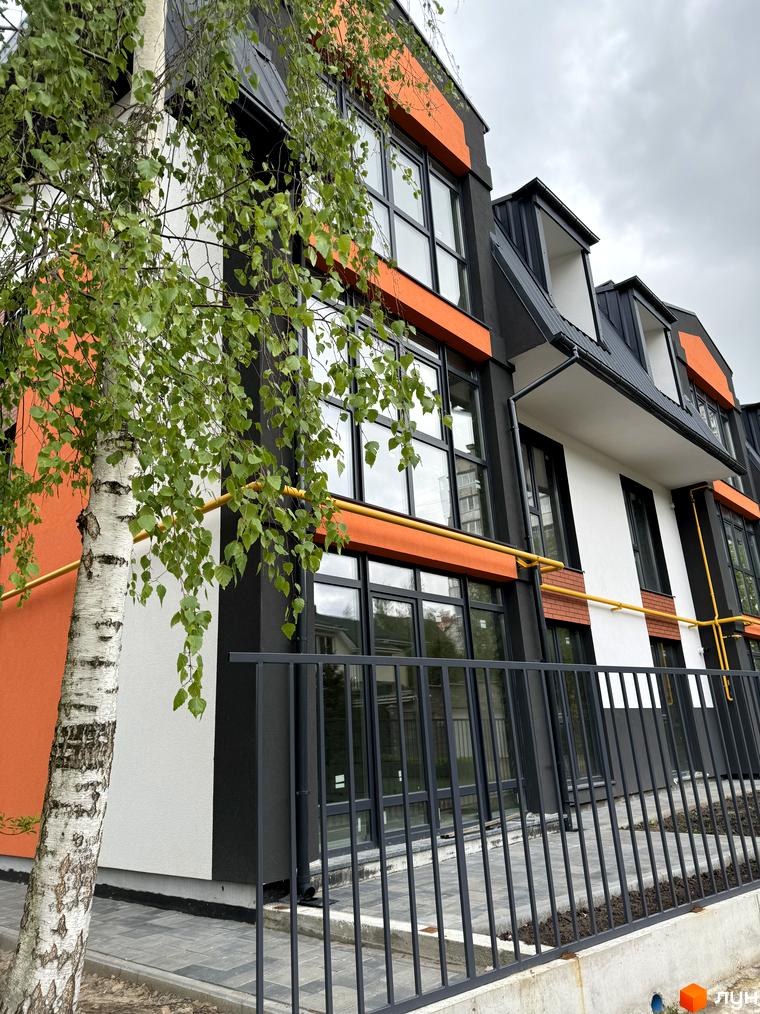Зовнішній вигляд новобудови Буковинська 20А з помаранчевими та сірими елементами фасаду та зеленим деревом перед будинком. Огорожа та бруківка на прибудинковій території.