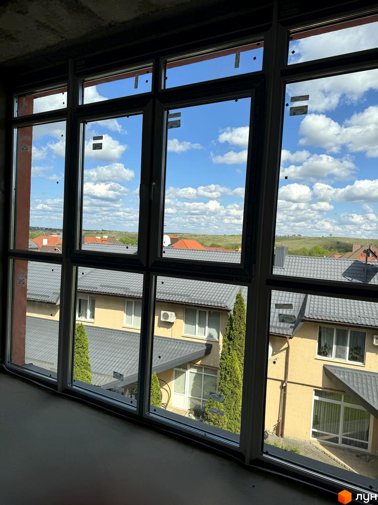 Вид з вікна на житловий комплекс Буковинська 20А з цегляними фасадами та сірими дахами. Навколо комплексу зелені насадження та блакитне небо з хмарами.