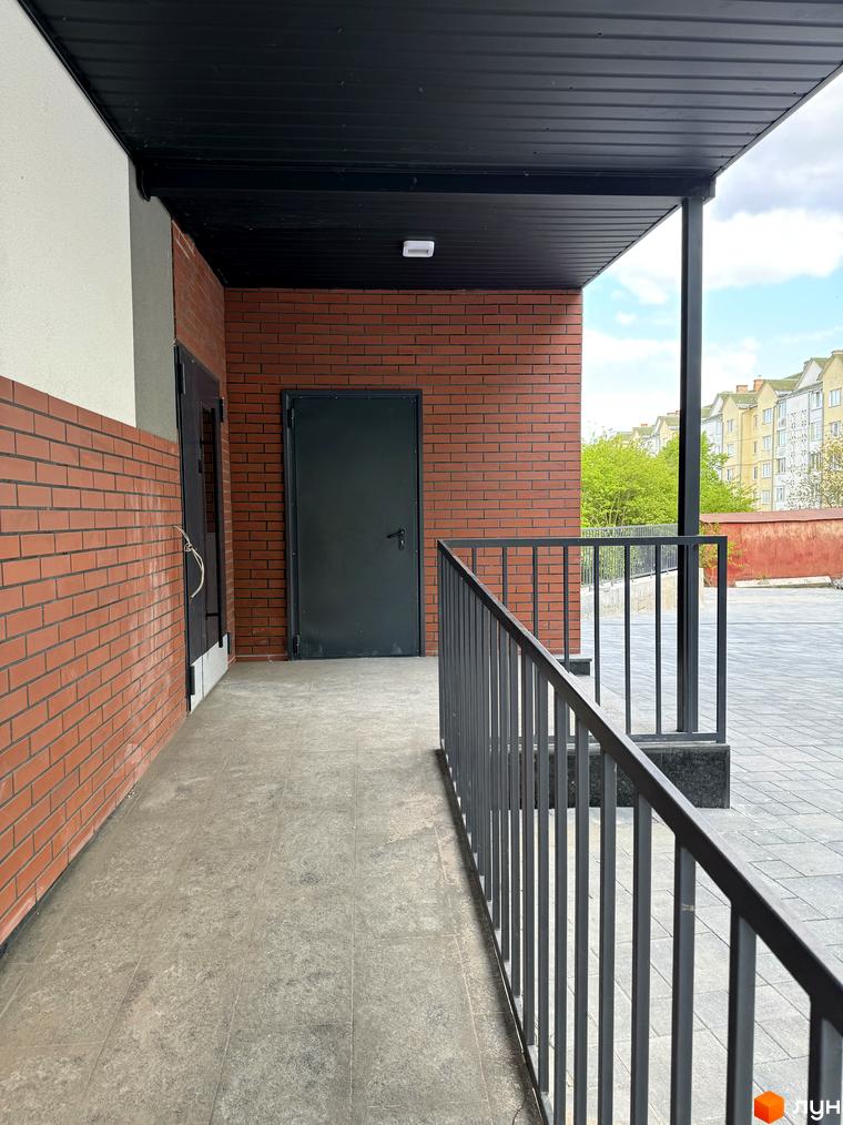 Зовнішній вигляд новобудови Буковинська 20А з цегляним фасадом та чорною покрівлею. Прибудинкова територія з вимощеним покриттям та металевою огорожею.