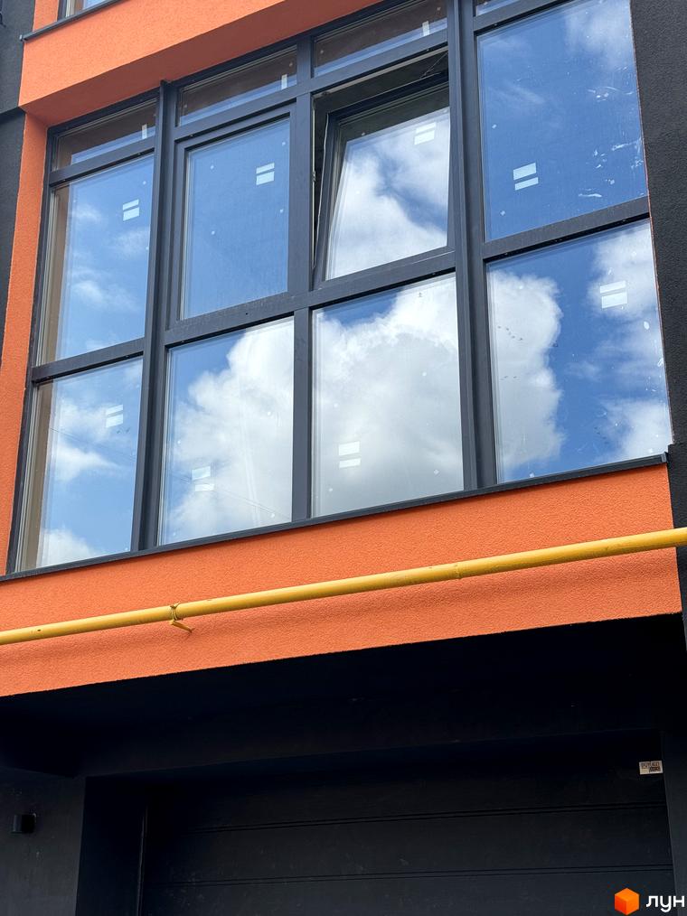 Фасад житлового комплексу Буковинська 20А з великими вікнами, що відбивають небо та хмари. Помаранчева штукатурка будівлі контрастує з темною нижньою частиною.
