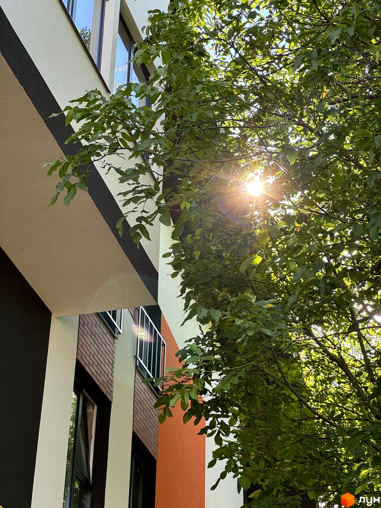 Фасад житлового комплексу Буковинська 20А з елементами цегляної кладки та оранжевими вставками, освітлений сонцем крізь зелене листя дерев.