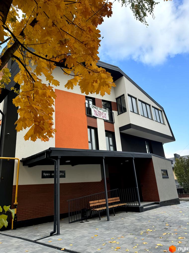 Житловий комплекс Буковинська 20А з фасадом білого, помаранчевого та коричневого кольору. Прибудинкова територія з лавкою та опалим листям.