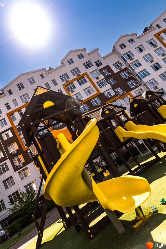 Житловий комплекс Orange Park. Біло-коричневі будівлі з численними вікнами та дитячий майданчик із жовтими гірками.
