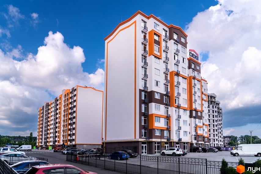 Житловий комплекс Orange Park з фасадами білого та коричневого кольорів з яскравими помаранчевими елементами. Навколо будівель розташований паркінг з автомобілями.