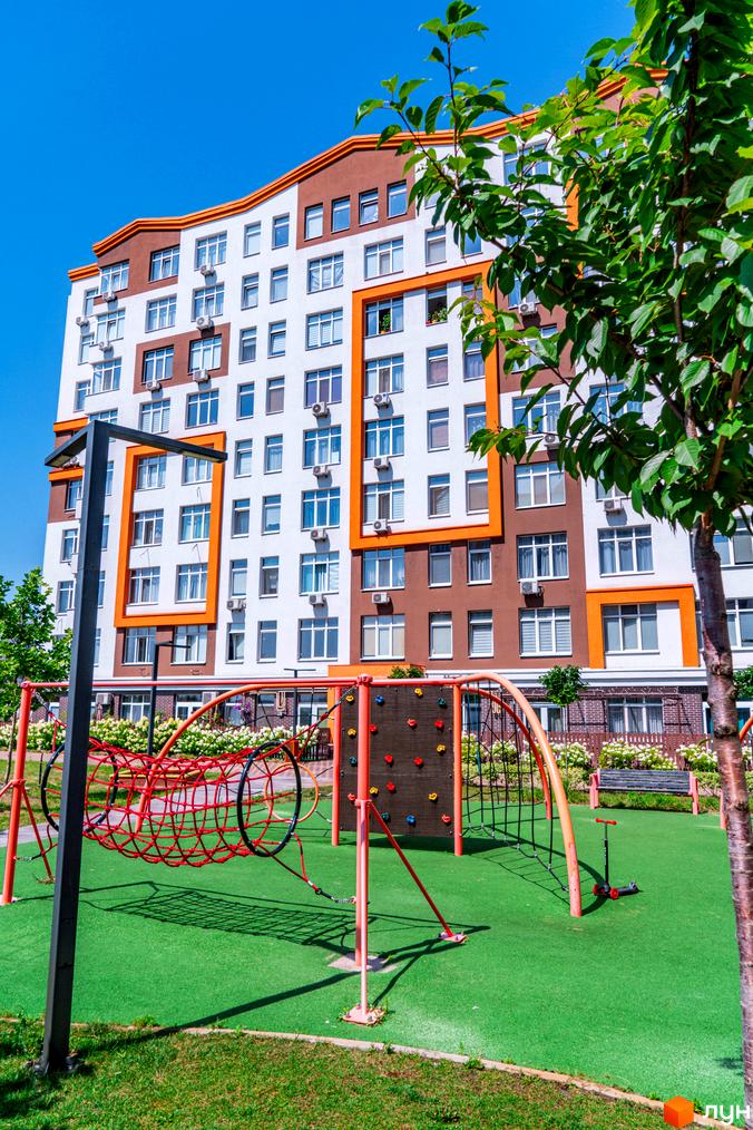 Житловий комплекс Orange Park: багатоповерховий будинок з біло-коричневим фасадом та оранжевими елементами, дитячий майданчик з червоними елементами та зеленою зоною.