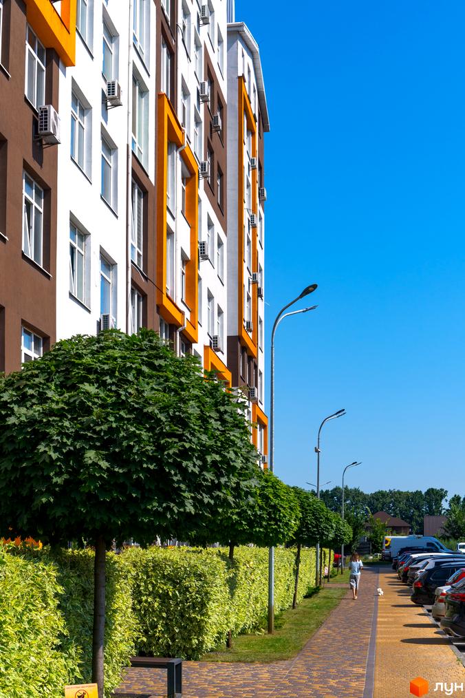 Житловий комплекс Orange Park з коричнево-білими фасадами та жовтими акцентами. Прибудинкова територія з доглянутими деревами, кущами та пішохідною доріжкою.