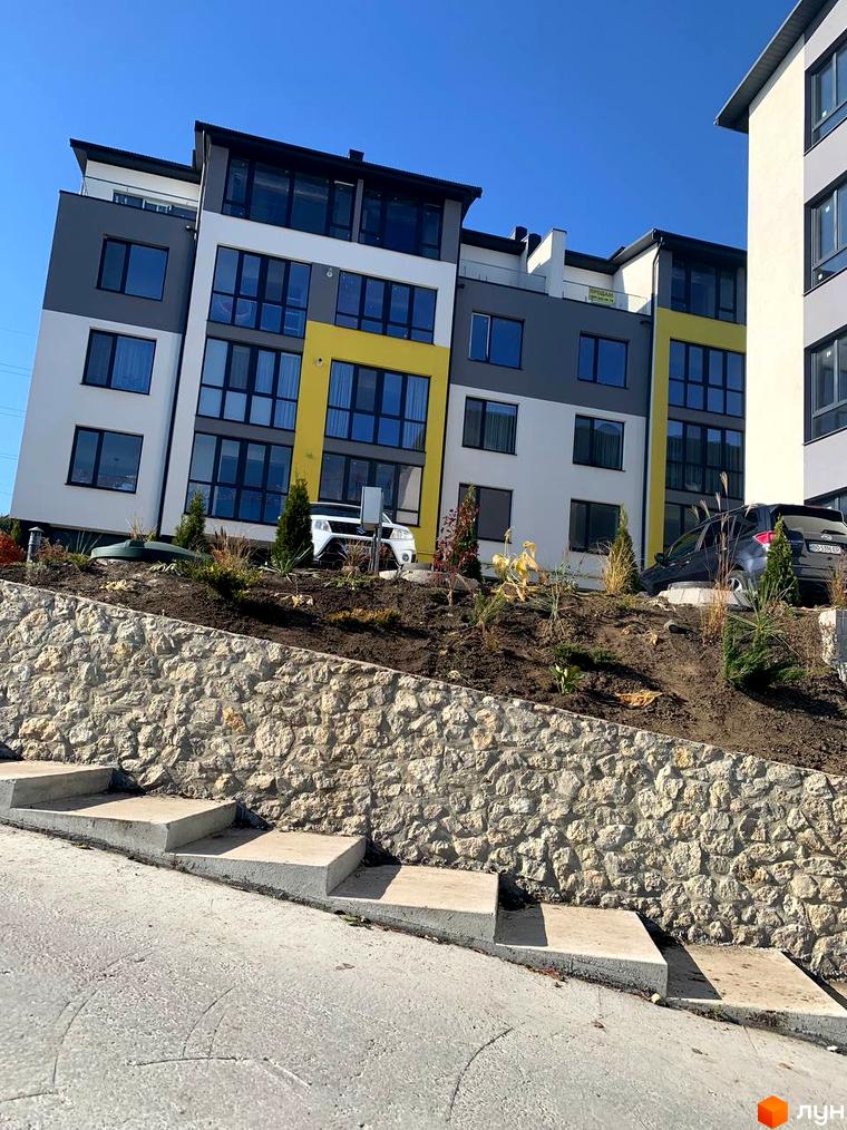 Багатоповерховий будинок з панорамними вікнами та жовтими вставками. Перед будинком кам'яна підпірна стінка та сходи.