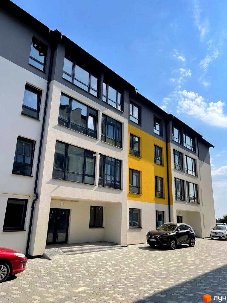 Фасад будинку Житловий комплекс ПАНОРАМА з білими та сірими стінами, великими вікнами та жовтою вставкою. На подвір'ї припарковані автомобілі, територія викладена плиткою.